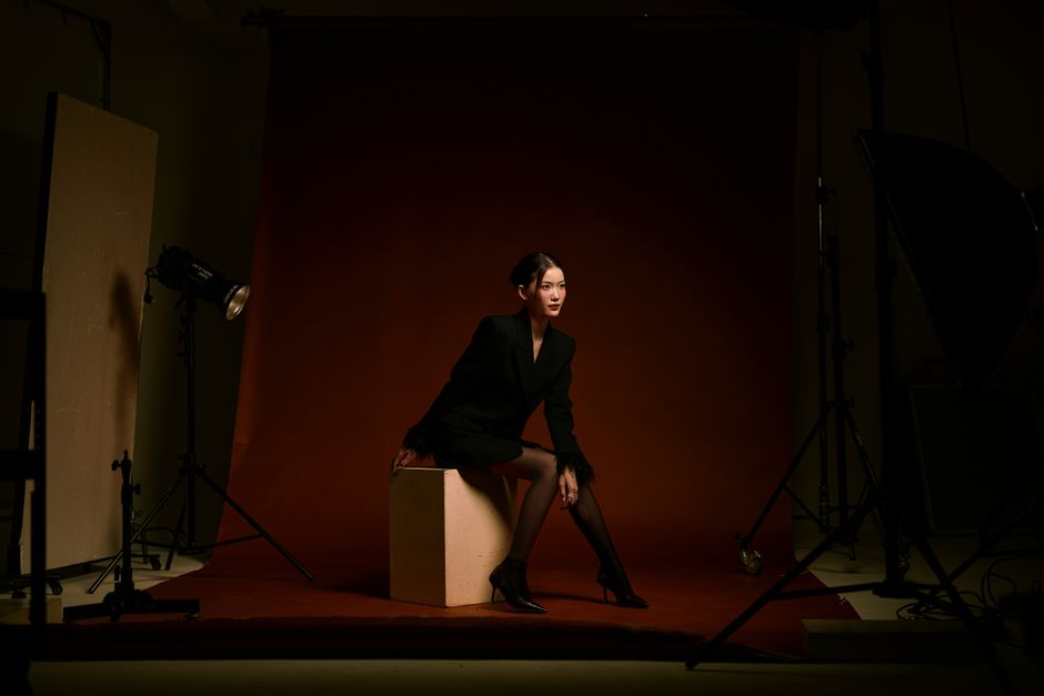 Stylish woman in black outfit sitting in studio with dramatic lighting.